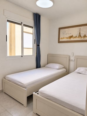 Une chambre avec deux lits simples, une fenêtre avec des rideaux, un luminaire fixé au mur et un tableau encadré représentant Paris.