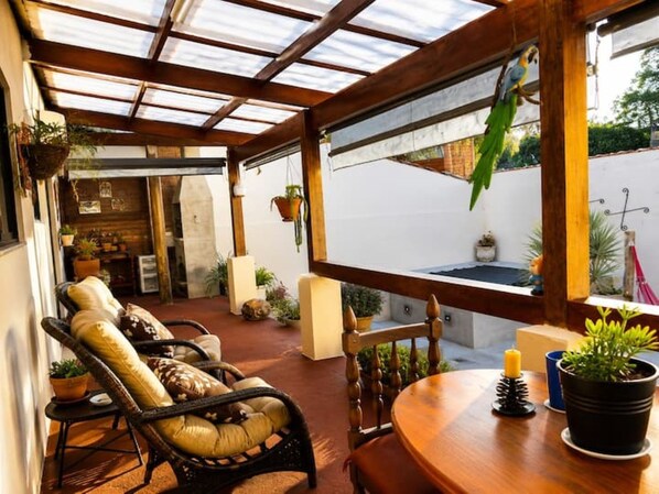 A covered outdoor seating area with a wooden table and chairs, surrounded by potted plants and a view of the street.