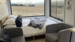 A bedroom with a large window, a bed with pillows, a bedside table, and two chairs.