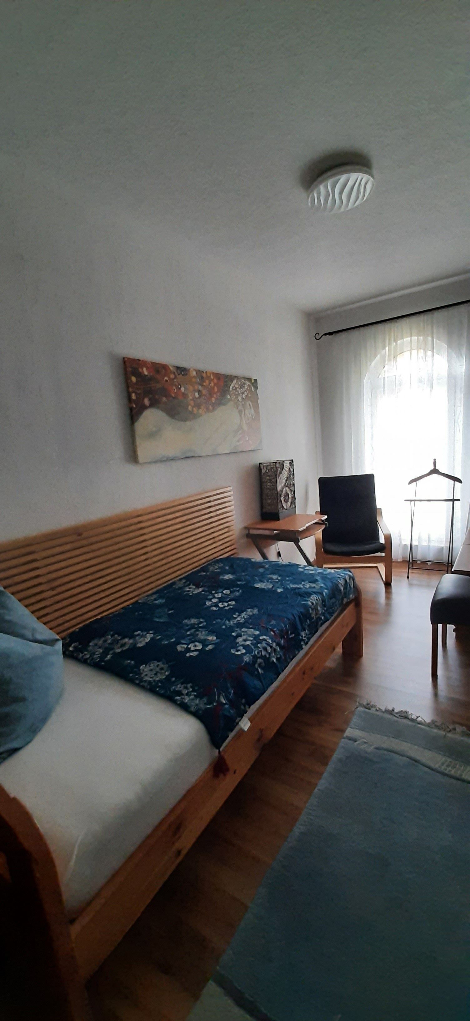 A bedroom with a wooden bed, a small table, a chair, and a painting on the wall.