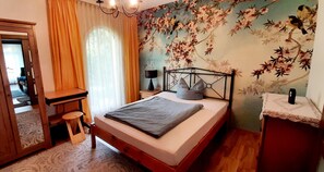 Un dormitori amb llit, escriptori, cadira, làmpada i un mur amb paper tapissat floral.
