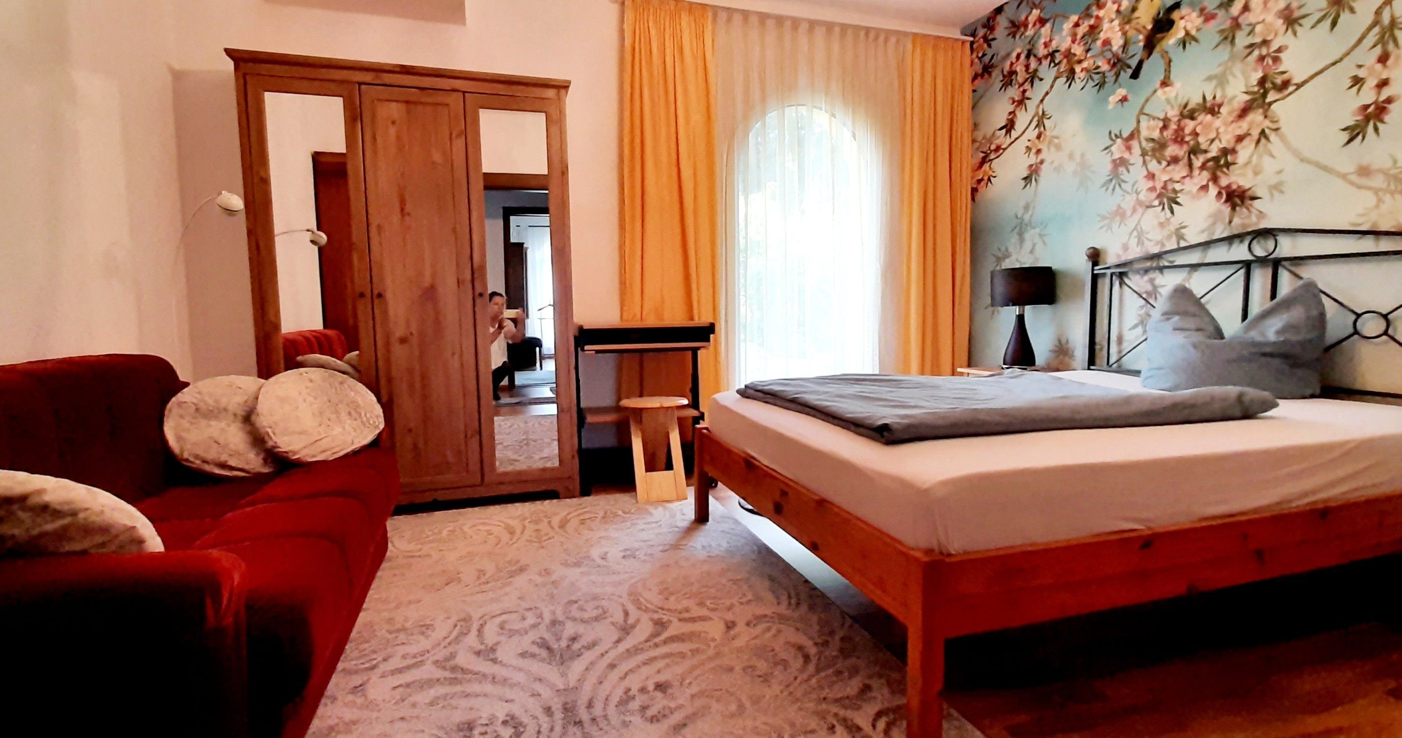 Ein Schlafzimmer mit einem hölzernen Bett, einem roten Sessel, einem kleinen Tisch und einer Tür zu einem anderen Raum.