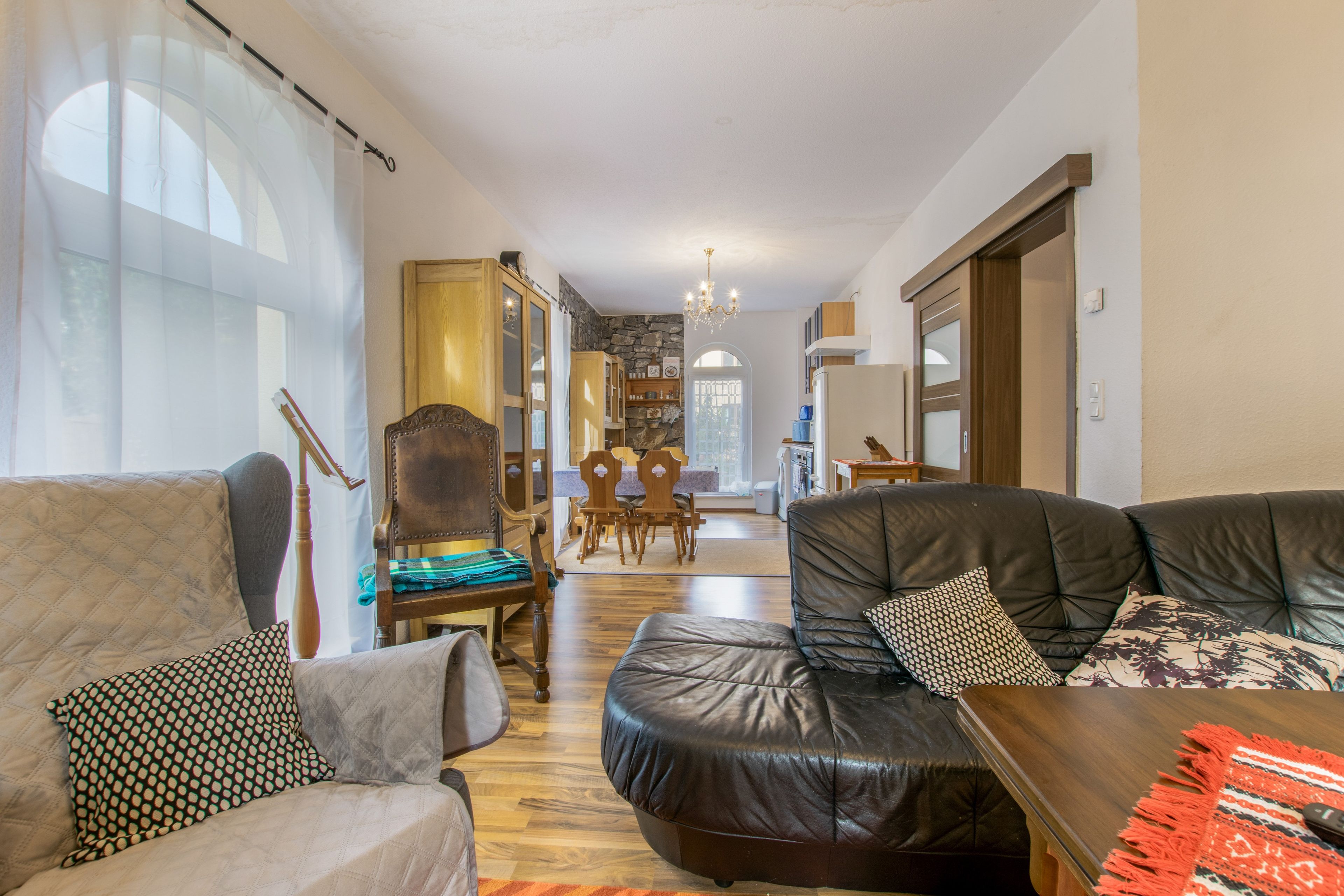 A spacious living room with a leather sofa, a wooden armchair, and a dining area with a stone wall and wooden furniture.