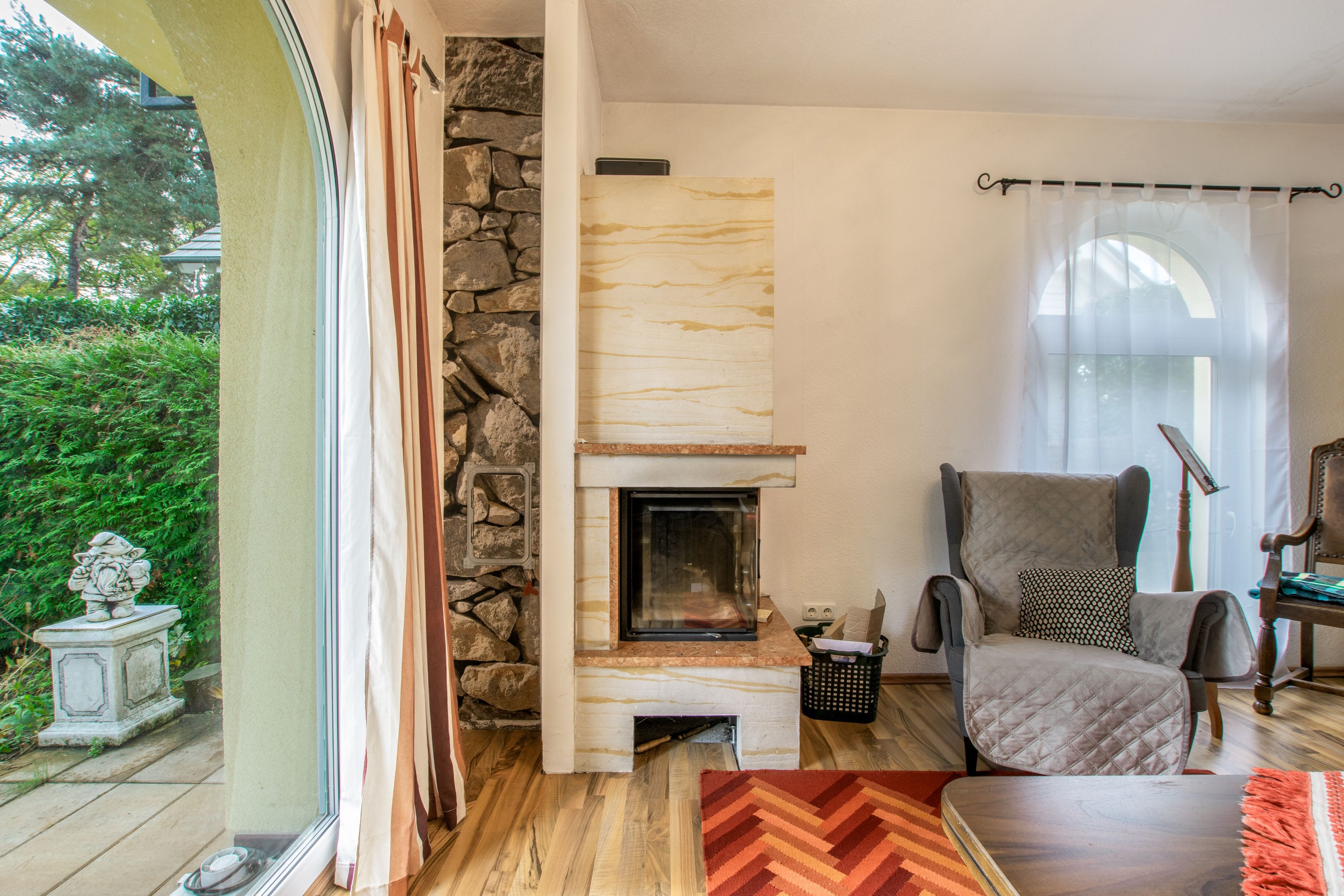 A cozy living room with a stone fireplace, a comfortable armchair, and a view of greenery outside.