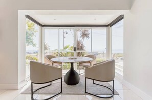 Ein Essbereich mit einem runden Tisch und vier Stühlen, großen Fenstern mit Blick auf Palmen und Gebäude, und einem hellen Interieur.