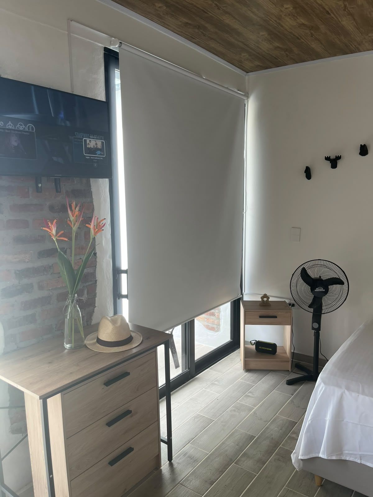 Une chambre à coucher avec un lit, un bureau avec des tiroirs, un ventilateur, une fenêtre donnant sur un beau paysage et un mur en briques.