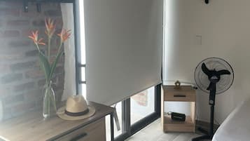 Une chambre à coucher avec un lit, un bureau avec des tiroirs, un ventilateur, une fenêtre donnant sur un beau paysage et un mur en briques.