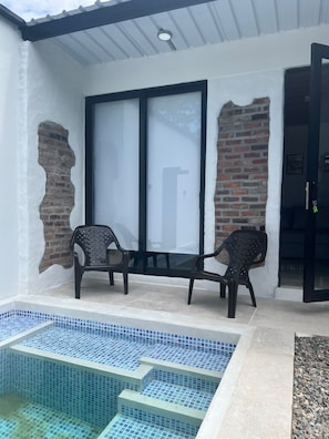 Uma área de estar coberta ao ar livre com duas cadeiras, piscina com azulejos e uma parede de tijolos.
