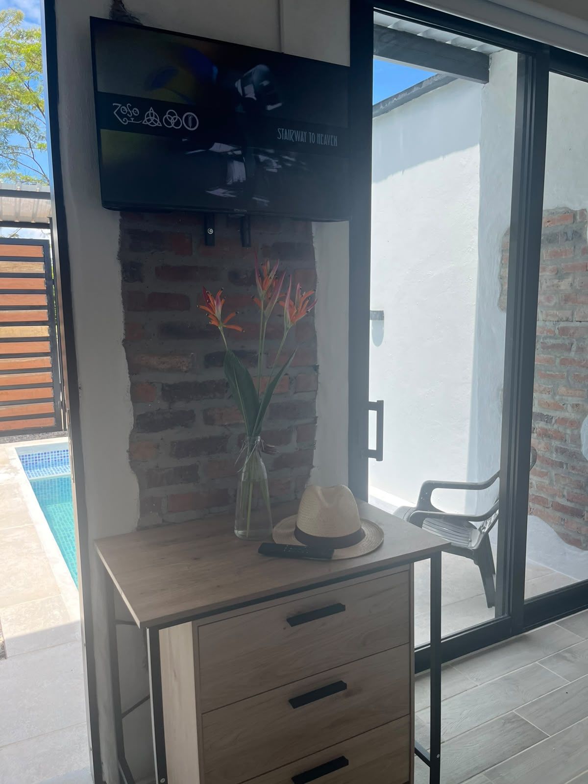 Habitación moderna con televisor montado en una pared de ladrillo, un escritorio de madera con cajones, un florero con flores y un sombrero.