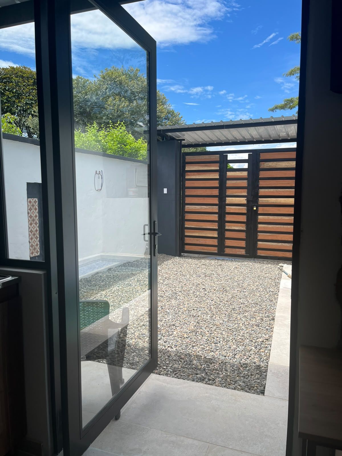 Un patio exterior moderno con puerta de vidrio que da a una zona de piscina, rodeado de grava y una valla de madera.