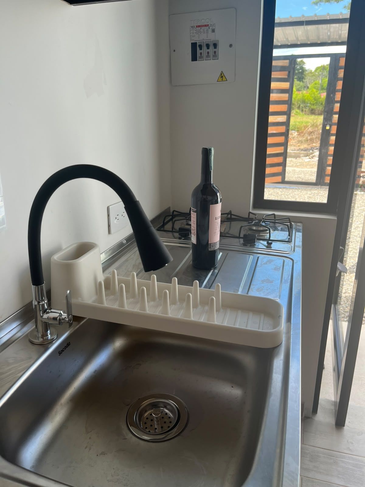 Una cocina con un lavamanos de acero inoxidable, una botella de vino y una ventana con vista a la vegetación.