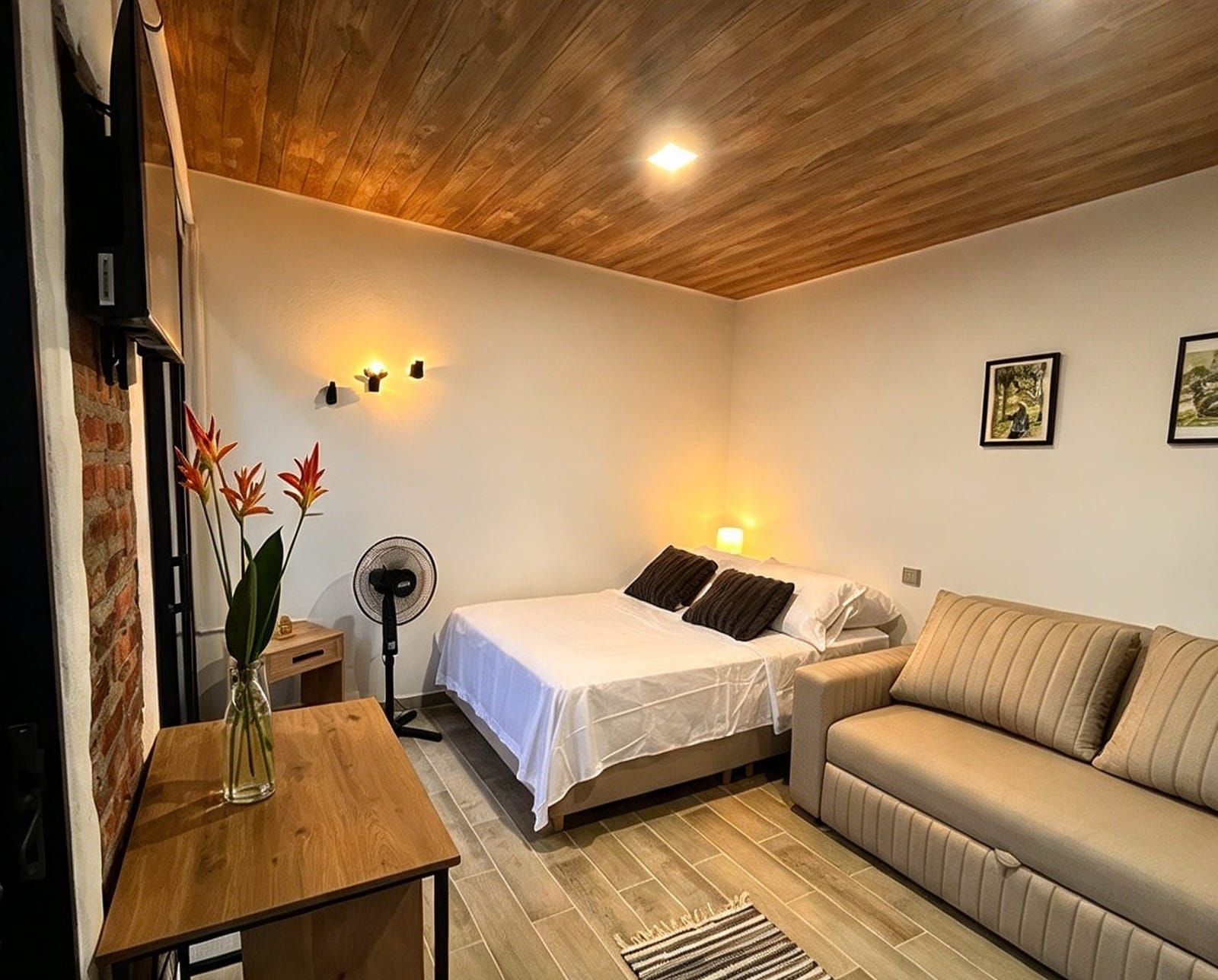 Una habitación de hotel con cama, sofá, ventilador, una mesita con un florero y una pared de ladrillo.