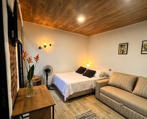Quarto de hotel com cama, sofá, ventoinha, mesinha com um vaso e parede de tijolos.