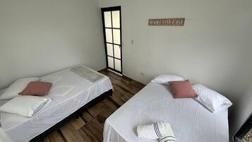 Una habitación con dos camas, cada una con sábanas blancas y una sola almohada. Hay una toalla sobre una de las camas. Un letrero en la pared dice 'BENDICE ESTA CASA'.