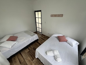 A room with two beds, each with white linens and a single pillow. A towel is placed on one bed. A sign on the wall reads 'BENDICE ESTA CASA.'