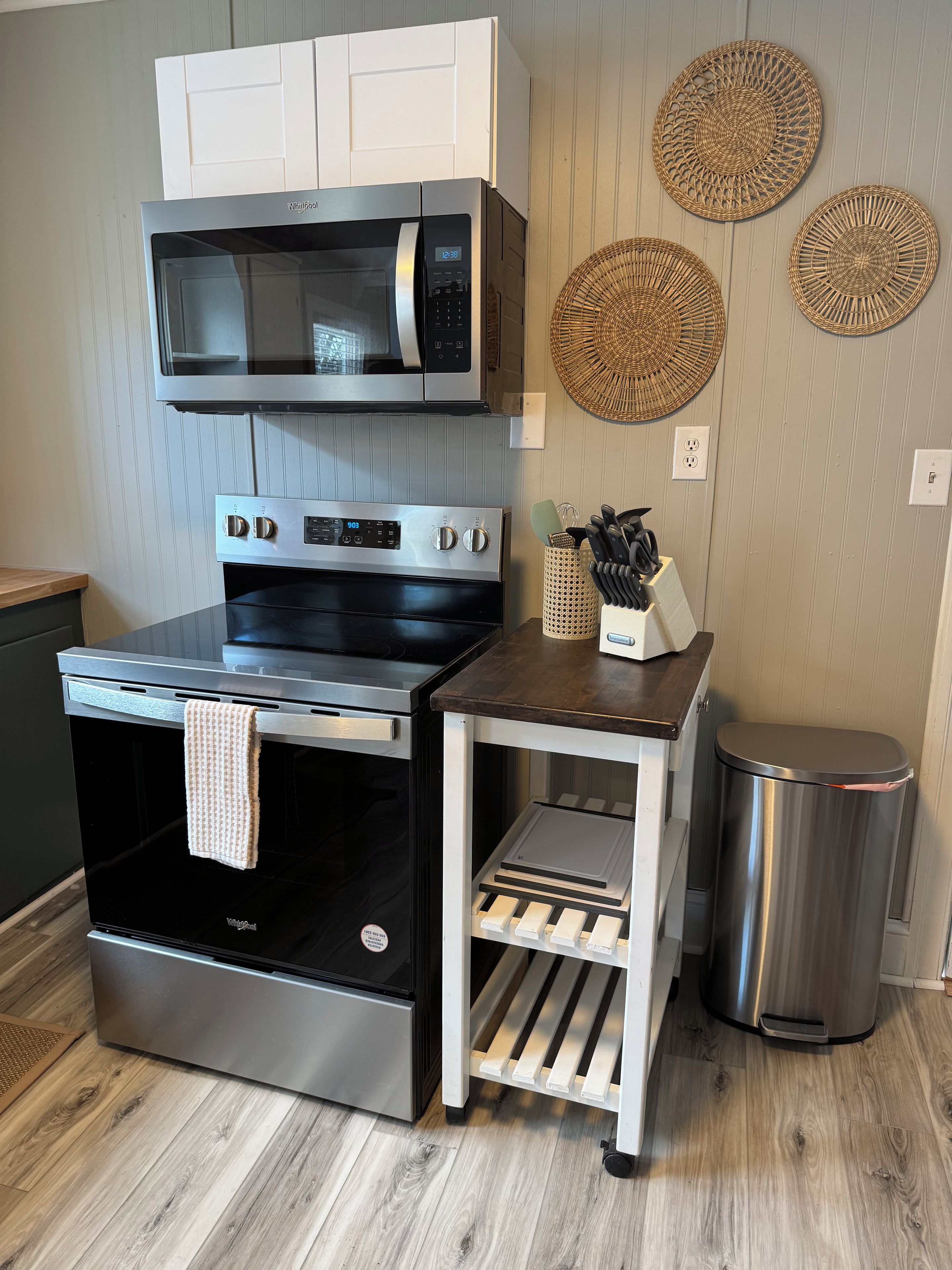 A kitchen with a stainless steel stove, microwave, and oven, a small wooden table with kitchen utensils, and a trash bin.
