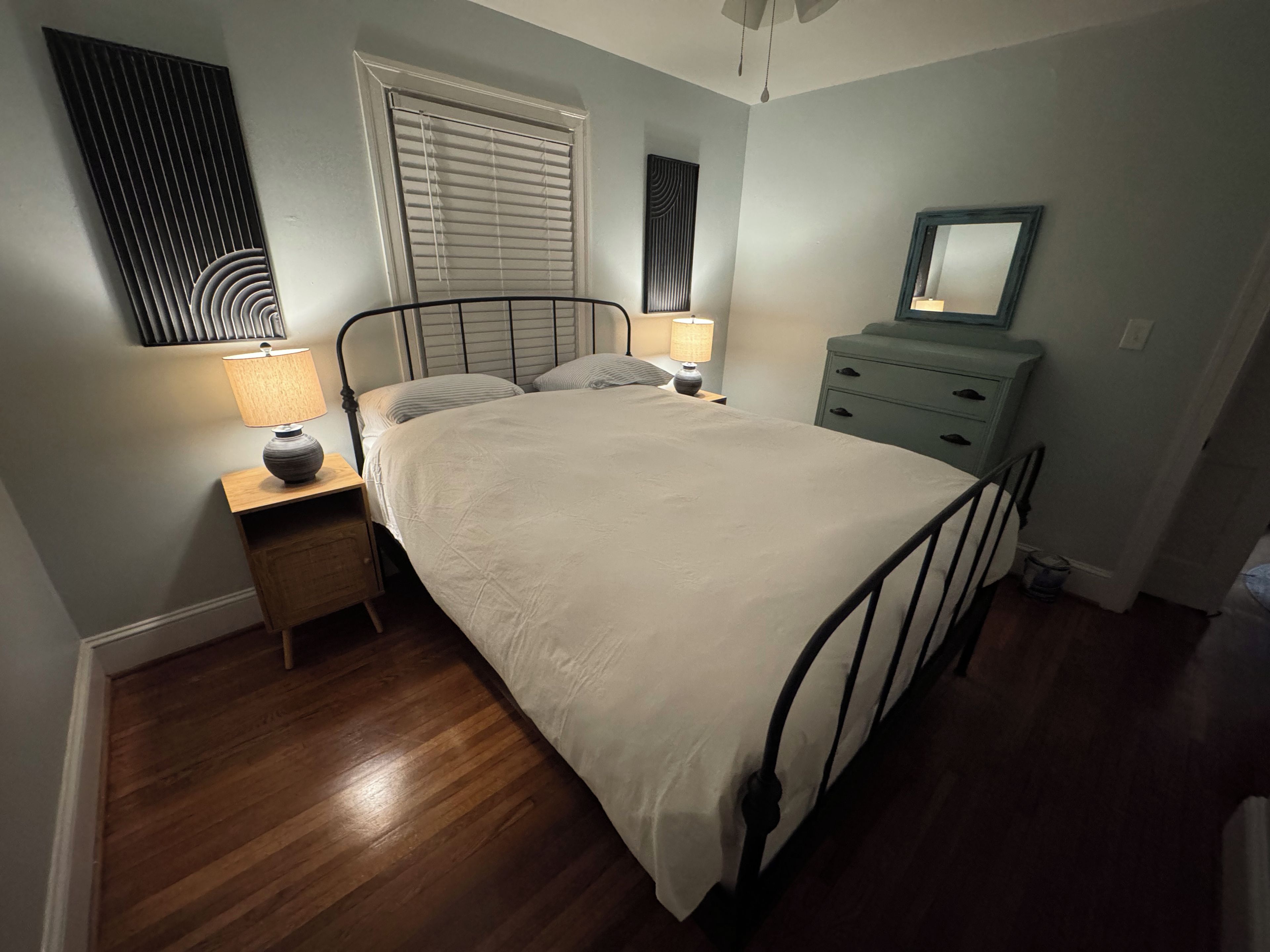 A bedroom with a bed, bedside tables, a dresser, a window with blinds, and a wall-mounted mirror.