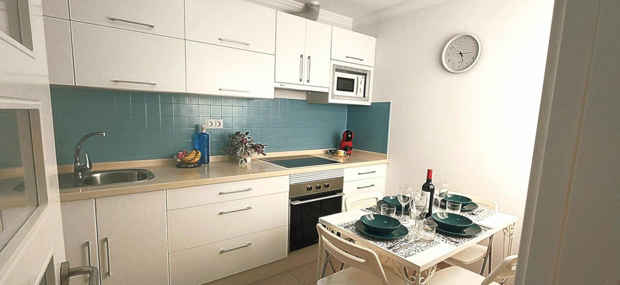 Een kleine keuken met witte kastjes, een magnetron, een spoelbak en een eethoek met een tafel gedekt voor twee personen.