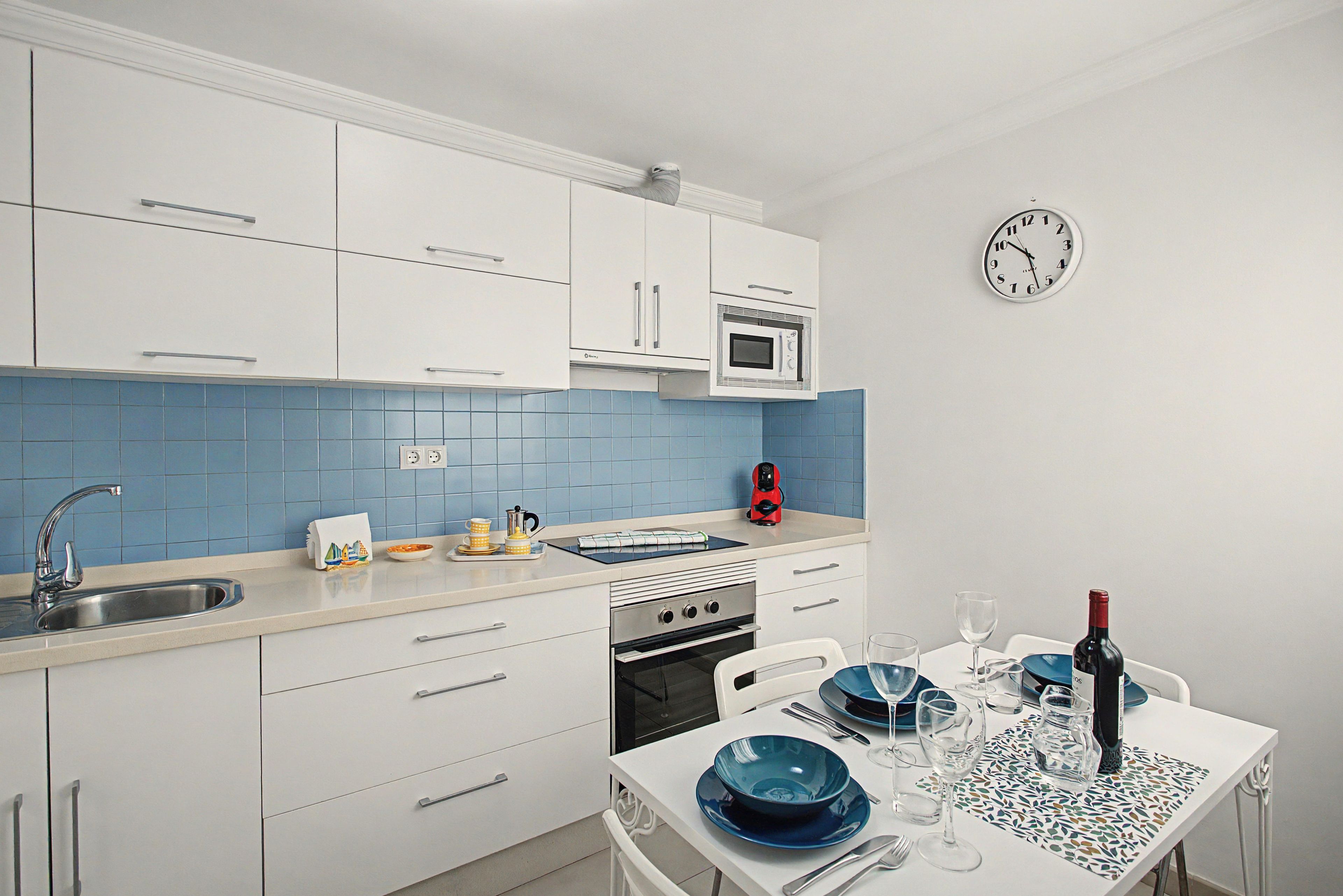 Una cocina moderna con armarios blancos, salpicadero de azulejos azules, microondas integrado y una zona de comedor con una mesa para dos.