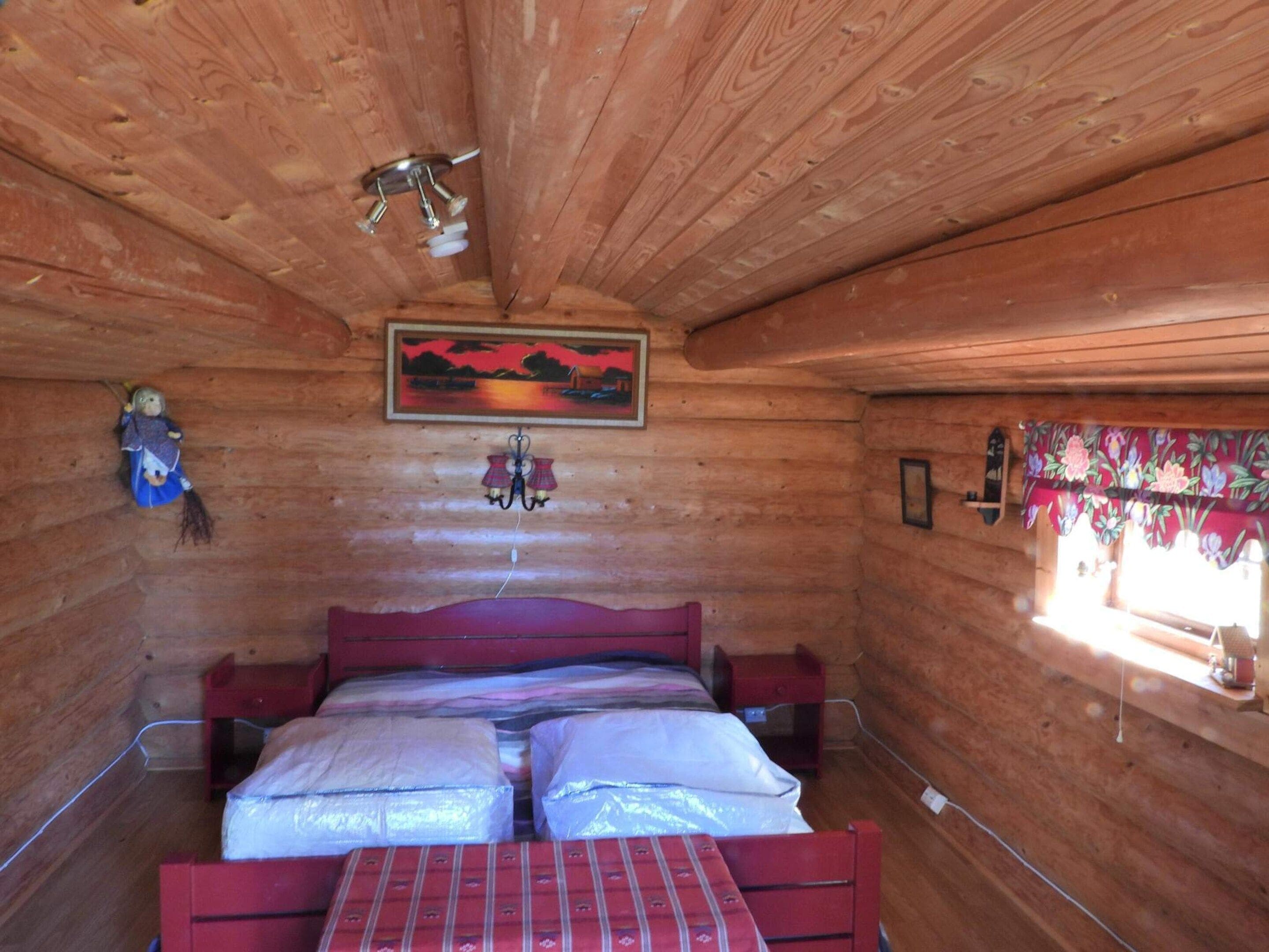 A wooden cabin room with a bed, a painting, a hanging lamp, and a window with curtains.