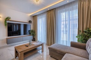 Una sala de estar moderna con un televisor de pantalla plana, una mesa de centro de madera, un sofá beige y un gran ventanal con cortinas translúcidas.