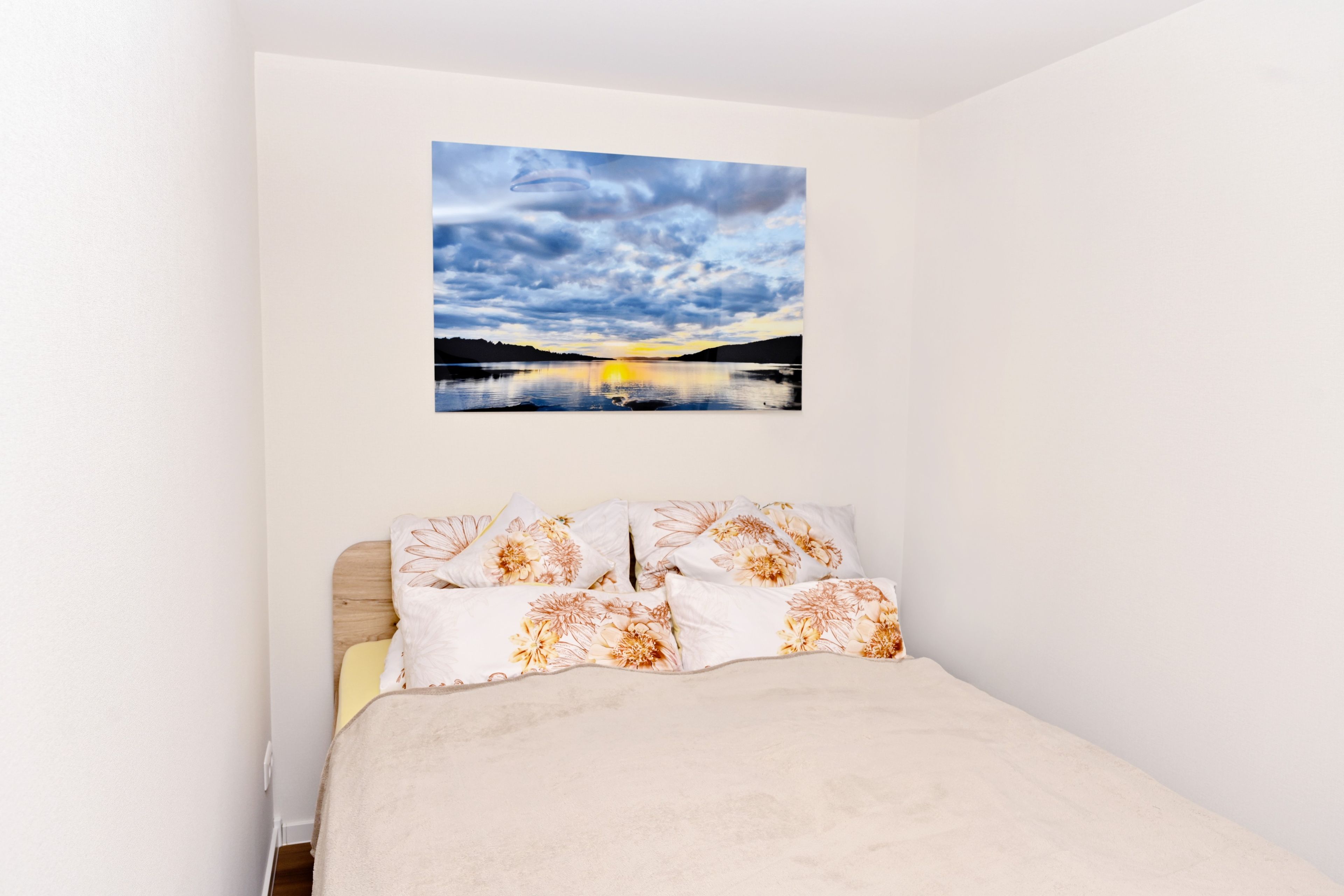 Ein Schlafzimmer mit einem Bett, einem Wandbild von einem Sonnenuntergang über Wasser und einer schlichten weißen Wand.
