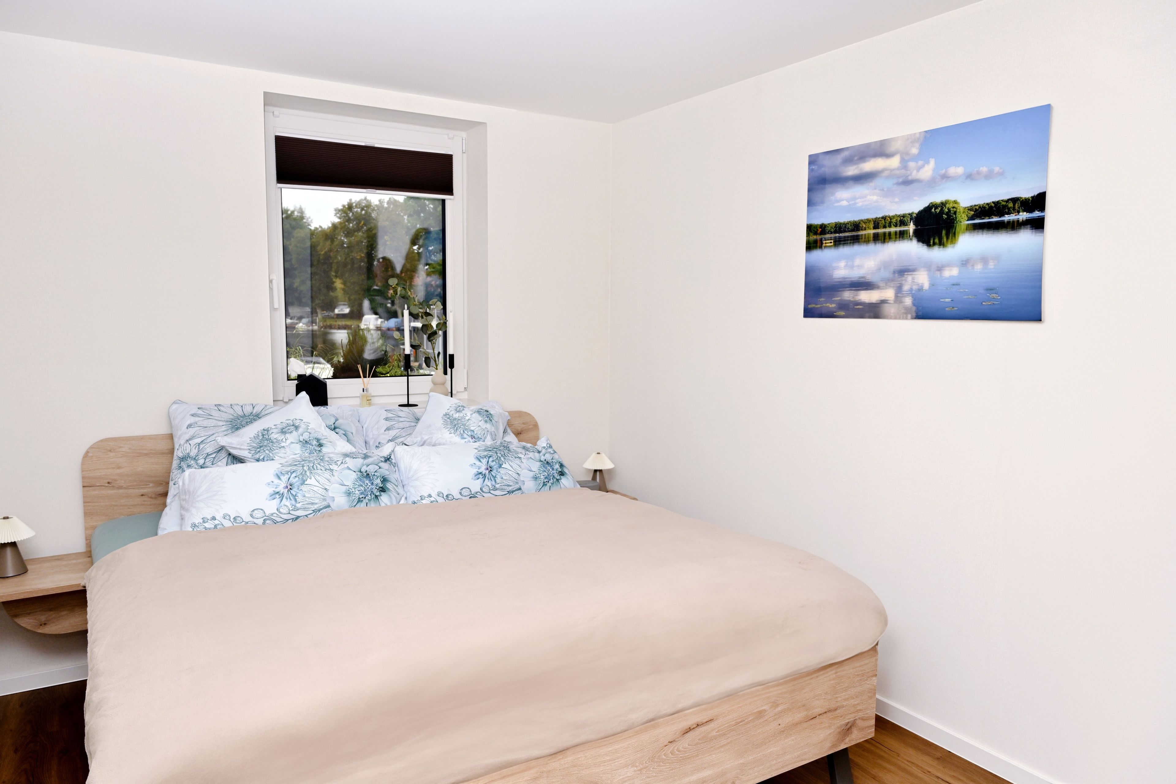 Ein Schlafzimmer mit Bett, Fenster, Nachttisch, Lampe und einem Wandbild.
