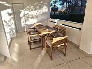 A sunlit patio with a wooden table and chairs, a sliding glass door, and a view of trees and sky. - Holiday Apartment With Air Conditioning (Risika)