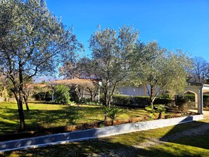 A residential area with a paved pathway, mature trees, and a building with a tiled roof. - Holiday Apartment With Air Conditioning (Risika)
