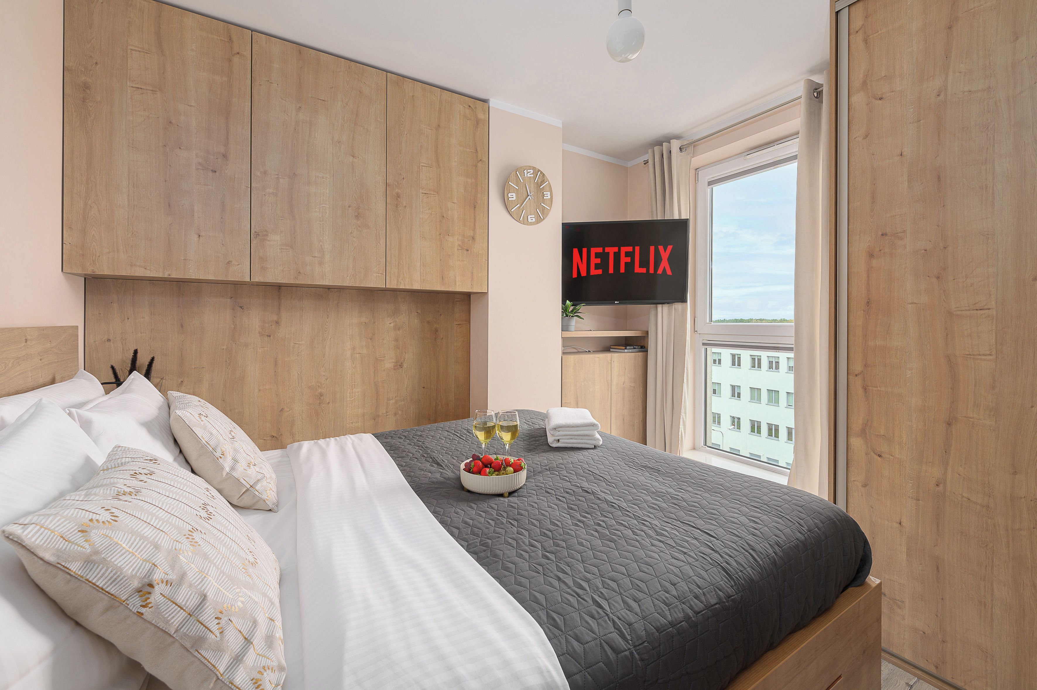 Un dormitorio moderno con cama, cabecero de madera, televisor de pared, un reloj y ventana con vistas.