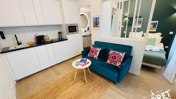 Un espacio habitable moderno y compacto con kitchenette, un sofá con cojines estampados y una cama con cabecera verde.