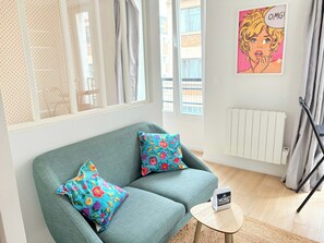 A room with a teal sofa, colorful floral cushions, a small round table with a sign, a radiator, and a window with curtains. - Moret Art Flats (Paris)