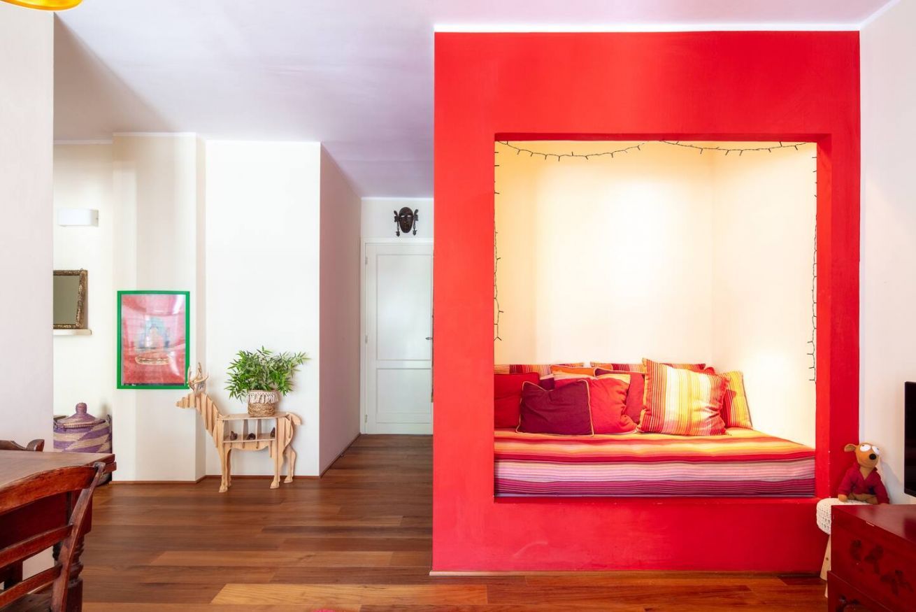 A room with a red accent wall, a wooden dining table, a chair, a small plant, and a sofa with colorful cushions.