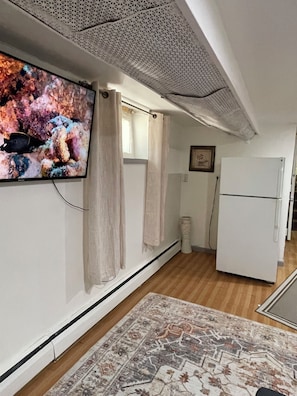 A room with a television mounted on the wall, a white refrigerator, a patterned rug, and a ceiling with a perforated design. - Cozy 1-bedroom house in lovely Jamaica with AC (Jamaica)