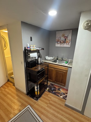 A compact kitchen with a microwave, kettle, and sink. There is a wooden cabinet and a small rug on the floor. - Cozy 1-bedroom house in lovely Jamaica with AC (Jamaica)