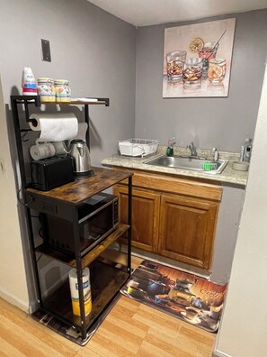 A compact kitchen with a microwave, toaster, and kettle on a wooden shelf, a sink with a faucet, and a small countertop.