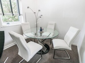 A modern room with a glass table surrounded by white chairs, a decorative metal plant, and a window with blinds. - 21 Ferrara Square (Swansea)