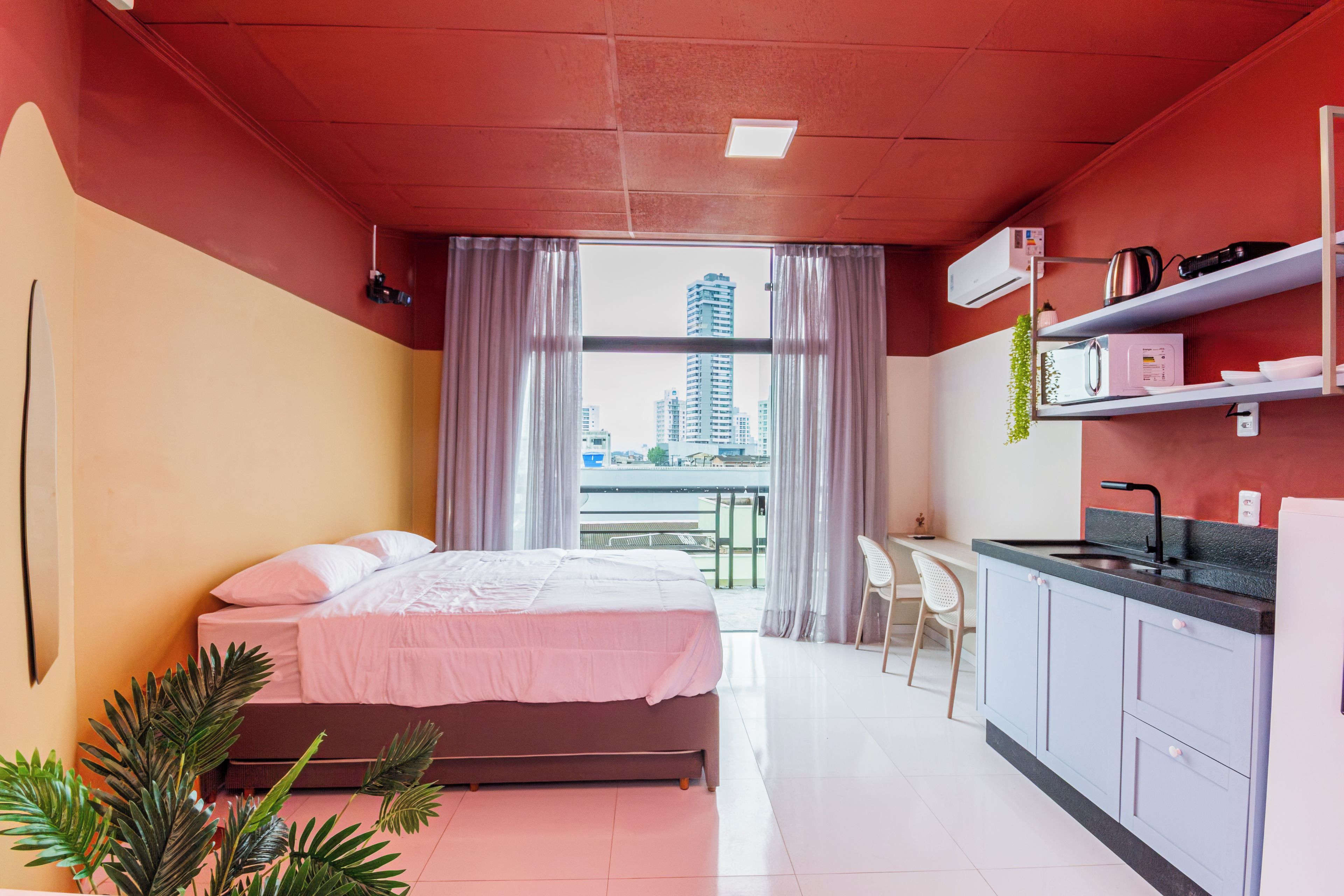 A modern hotel room with a bed, a small kitchenette, and a view of the city.