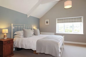 A bedroom with a white bed, a nightstand with a lamp, a window with blinds, and a picture on the wall. - Bryher Cottage (Newquay)