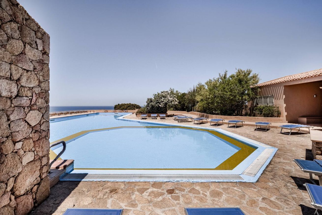 Ein Swimmingpool mit einer Steinsäule an einer Seite, umgeben von Liegestühlen und einem Gebäude mit einem Ziegeldach.