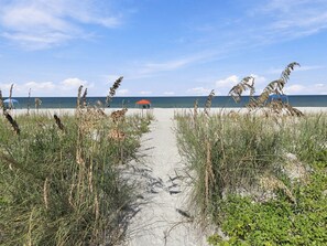 Ein Strandweg führt zu einem roten Sonnenschirm, im Vordergrund Sand und Seegras.