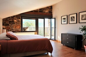 A bedroom with a stone wall, a bed with a red bedspread, a black dresser, and a balcony with a view. - Casa de vacaciones 'Casa Yañez' con vistas a la montaña, terraza y Wi-Fi (Chantada)