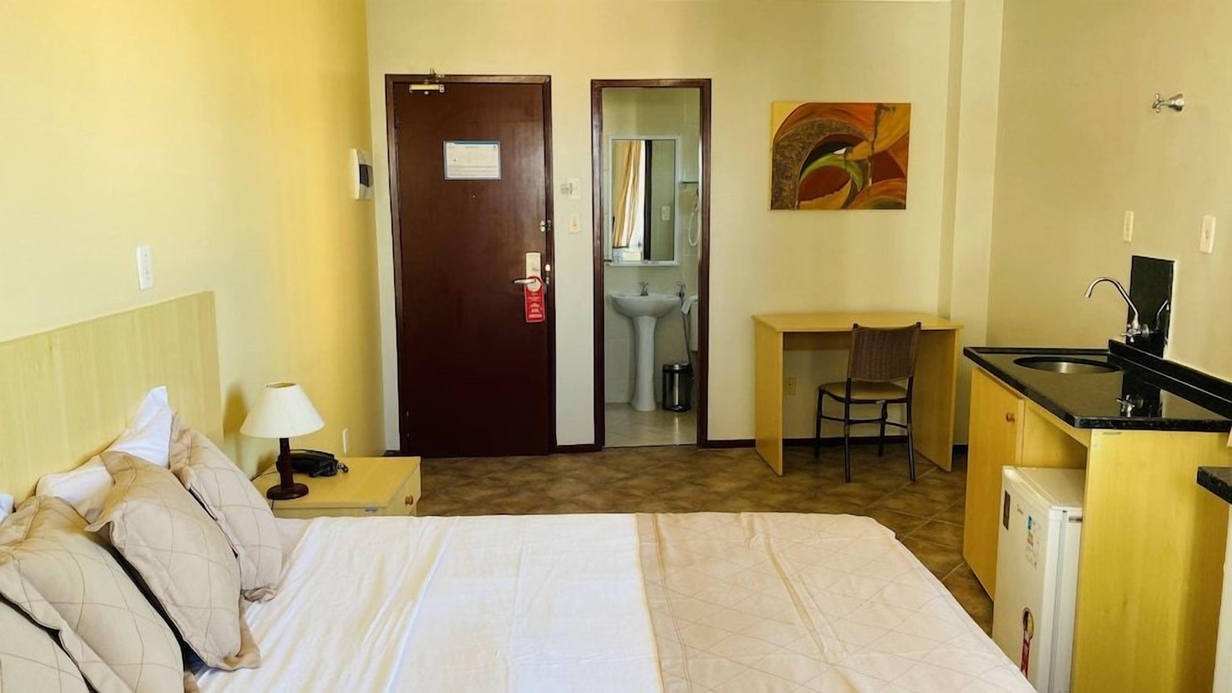 A hotel room with a bed, a desk, a chair, a sink, and a refrigerator.