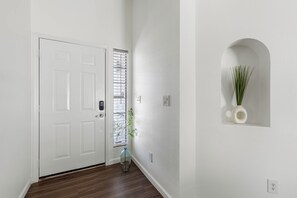 A hallway with a white door, a window with blinds, a potted plant, and a wall-mounted light fixture. - 3 Bedroom/ Private Fenced Backyard /Desert Style (Gilbert)
