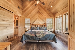 Ein Schlafzimmer einer Holzhütte mit einem Bett, einem Deckenventilator und großen Fenstern.