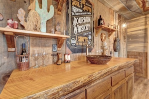 A rustic bar area with wooden shelves, a whiskey bottle, glasses, and decorative items.