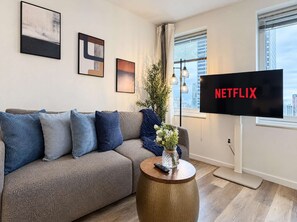 En stue med en grå sofa, blå puder, et træsofabord med en vase, og et fjernsyn der viser 'NETFLIX'.