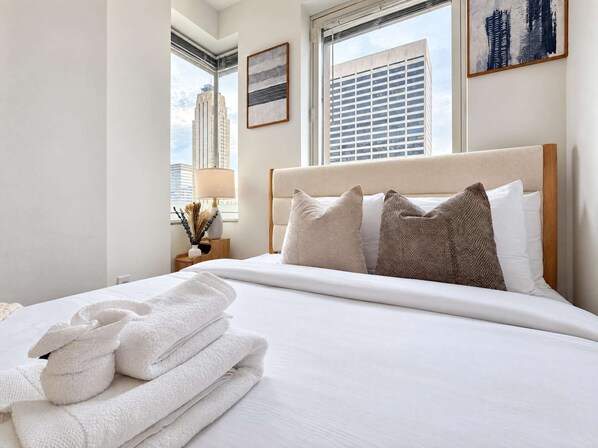 A hotel room with a large bed, a nightstand with a lamp, and a window with a city view.
