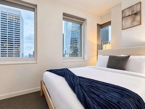Ein Hotelzimmer mit einem großen Bett, zwei Fenstern, Blick auf hohe Gebäude und einem Gemälde an der Wand.