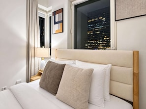 A hotel room with a bed, pillows, a nightstand, a lamp, a window with a city view, and a framed picture on the wall.